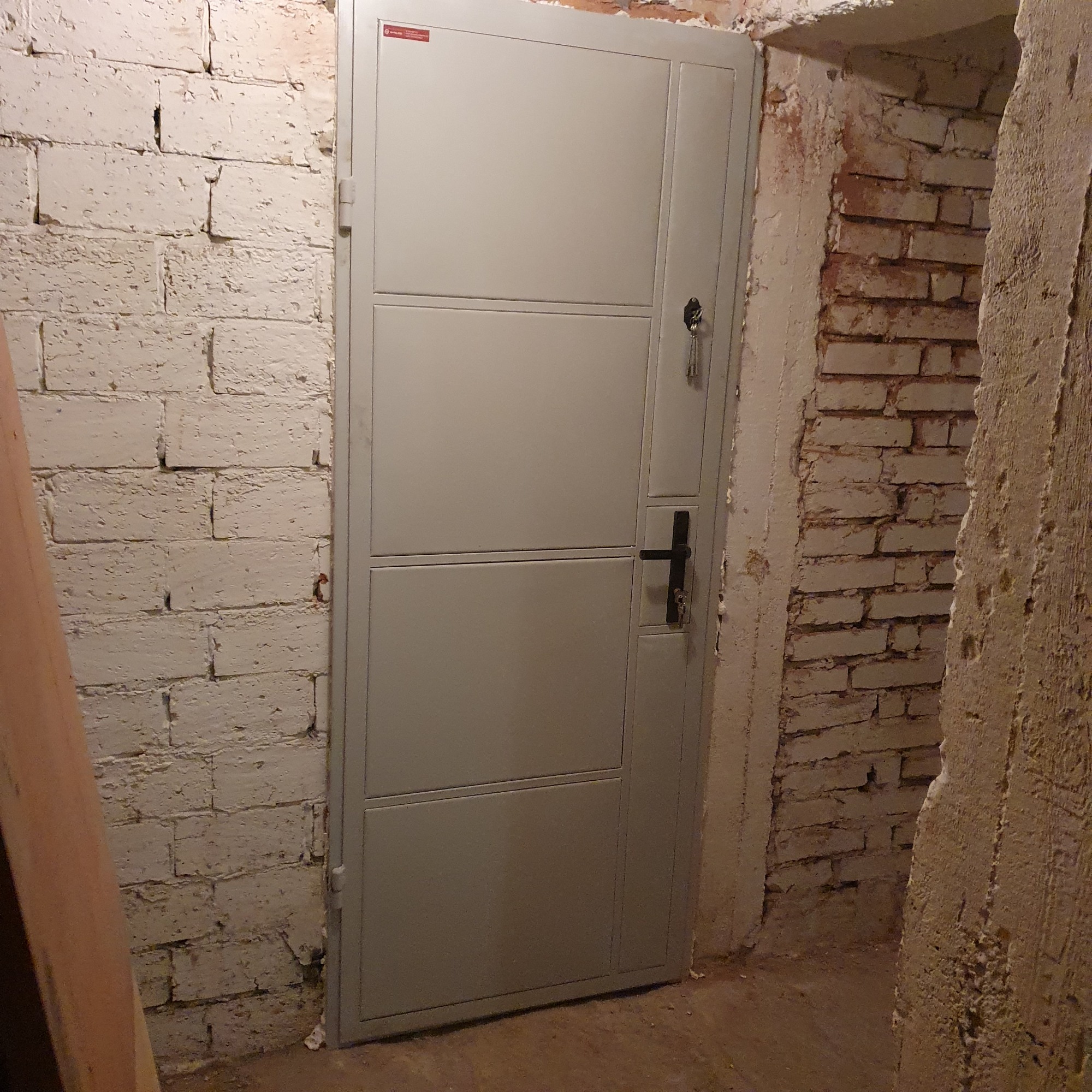 Szare, metalowe drzwi z czarną klamką i kluczami w zamku, zamontowane w surowym, ceglanym murze.