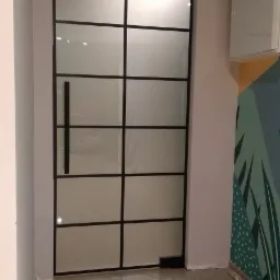 Matowe, przeszklone drzwi wewnętrzne z czarną ramą i podłużnym uchwytem, widoczne na tle wzorzystej ściany i jasnej podłogi.