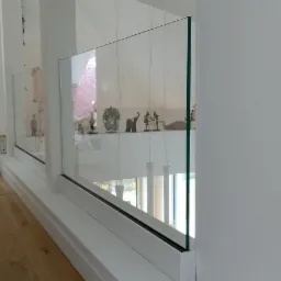 Nowoczesna szklana barierka z minimalistycznymi, białymi ramami i dekoracyjnymi figurkami na półce, widok z boku na drewnianą podłogę.