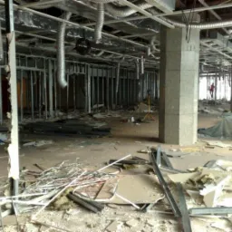 Wnętrze budynku w trakcie demontażu ścianek działowych, widoczne metalowe profile, gruz, odsłonięta instalacja pod sufitem, filary z szarego kamienia oraz częściowo zabezpieczone folią elementy...