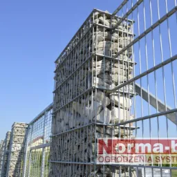 Ogrodzenie gabionowe z siatki i kamienia na tle błękitnego nieba. Widoczny fragment ogrodzenia z bramą i logo producenta.