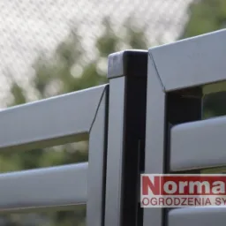 Fragment grafitowego ogrodzenia panelowego z logo firmy Norma-bud na tle zieleni i siatki. Widoczne detale łączeń i estetyczne wykończenie.