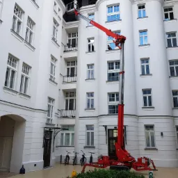 Czerwony podnośnik koszowy ustawiony na dziedzińcu kamienicy z białymi ścianami i licznymi oknami, kosz podnośnika sięga do górnych kondygnacji budynku.