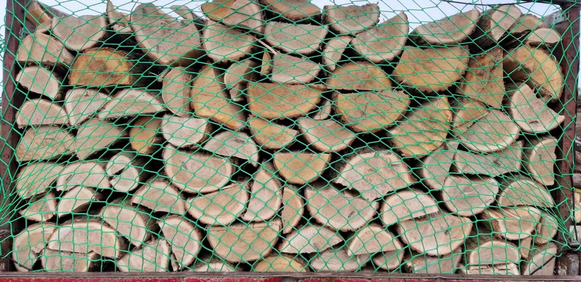 Rzędami ułożone, pocięte kawałki drewna opałowego zabezpieczone zieloną siatką transportową w skrzyni ładunkowej.