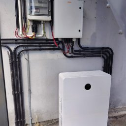 ECONOWATOR - Inwerter fotowoltaiczny KENO, rozdzielnica elektryczna z bezpiecznikami oraz duża biała skrzynka, zamontowane na szarej ścianie. Widoczne czarne peszle i kolorowe przewody.
