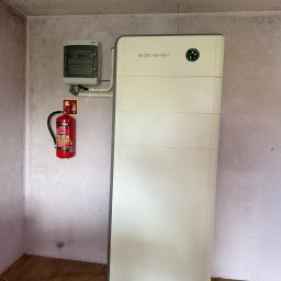 ECONOWATOR - Zainstalowany biały magazyn energii SIDENERGY z zielonymi diodami, połączony z rozdzielnicą elektryczną i gaśnicą, w pomieszczeniu z drewnianą podłogą.