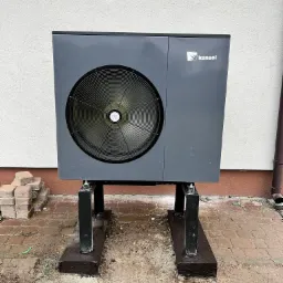 Szara pompa ciepła Kensol zamontowana na czarnych, metalowych wspornikach, ustawiona na drewnianych podstawach na błotnistym podłożu z kostki brukowej, przy ścianie budynku z rynną.