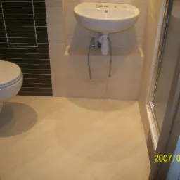 Jasna łazienka z beżową glazurą terakotą na podłodze, białą umywalką bez baterii i toaletą z czarnymi akcentami na ścianie, widok z wejścia do kabiny prysznicowej.