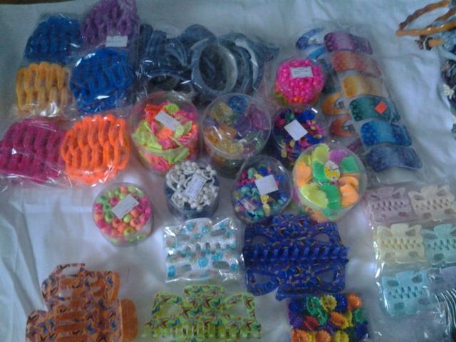 Różnorodny asortyment sztucznej biżuterii: kolorowe gumki do włosów w siateczkach, spinki do włosów w przezroczystych opakowaniach oraz klamry do włosów z wzorami, ułożone na białym materiale.