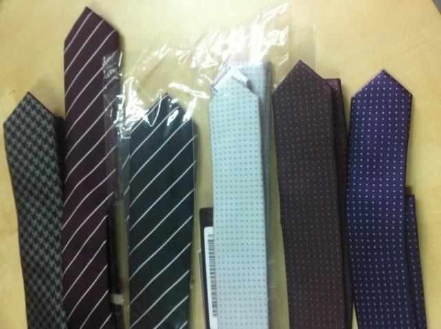 Siedem krawatów w różnych wzorach i kolorach ułożonych na jasnym blacie, niektóre zapakowane w folię, widoczny kod kreskowy.
