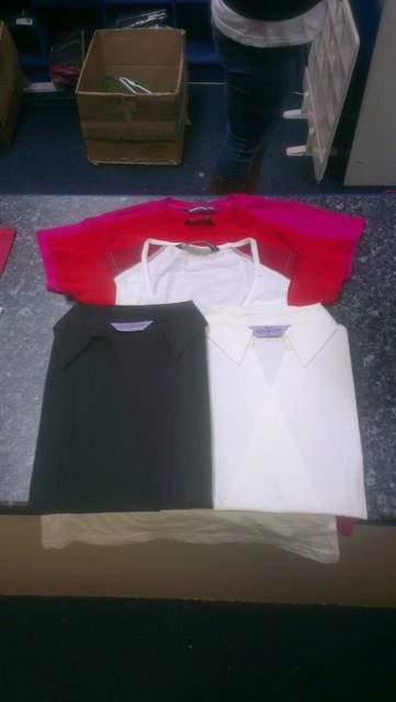 Ułożone na blacie koszulki: czerwona, biała na ramiączkach, czarna koszula z kołnierzykiem i biała koszula z kołnierzykiem, w tle kartonowe pudełko i fragment osoby w jeansach.