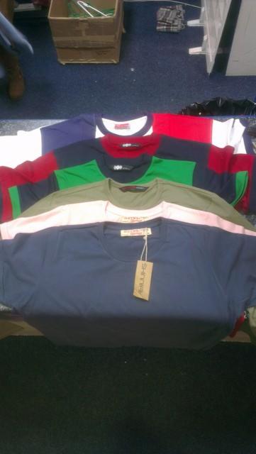 Stos ułożonych koszulek damskich, męskich i dziecięcych w różnych kolorach, z widoczną metką na dolnej koszulce.