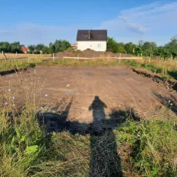 Przygotowany teren pod budowę domu, widoczny fundament, w tle biały dom z ciemnym dachem, cień fotografa na pierwszym planie.