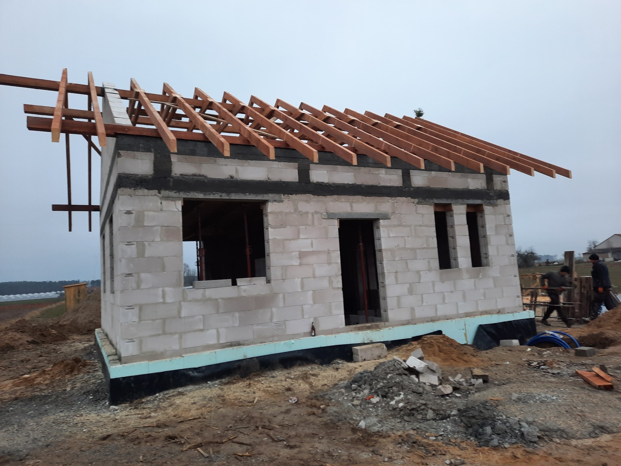 Surowy budynek z białych bloczków, z widoczną konstrukcją drewnianą dachu, ocieplony fundament, w tle krajobraz z polami i szklarniami.