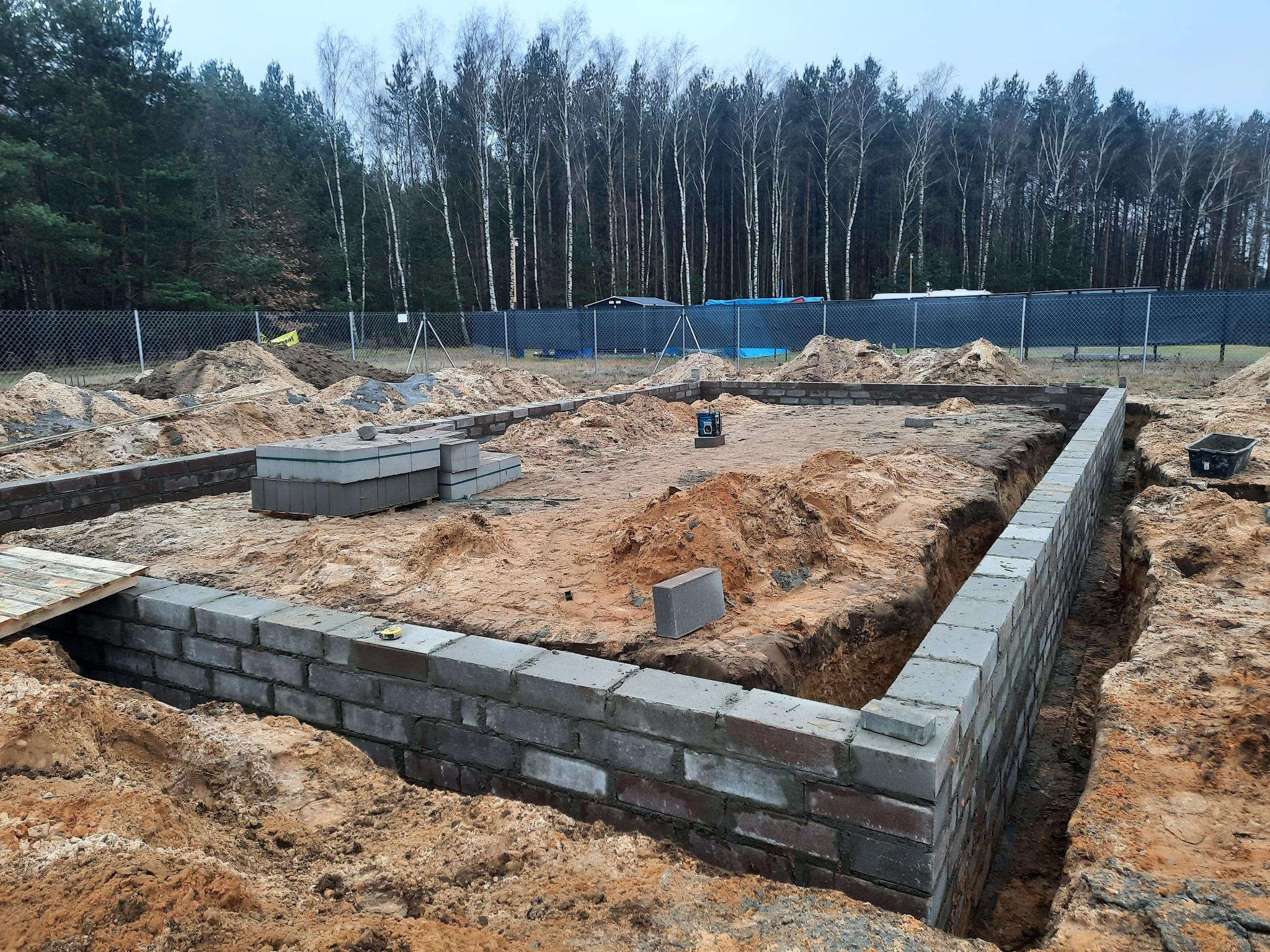 Wykop pod fundamenty domu murowanego z częściowo wzniesionymi ścianami fundamentowymi z szarej cegły, widoczny laser krzyżowy, w tle las i ogrodzenie.
