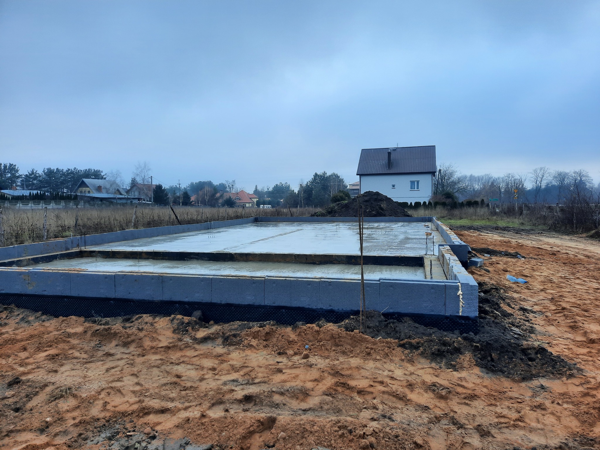 Wylana płyta fundamentowa z izolacją, widok na budowę domu w pochmurny dzień, w tle zabudowa jednorodzinna.