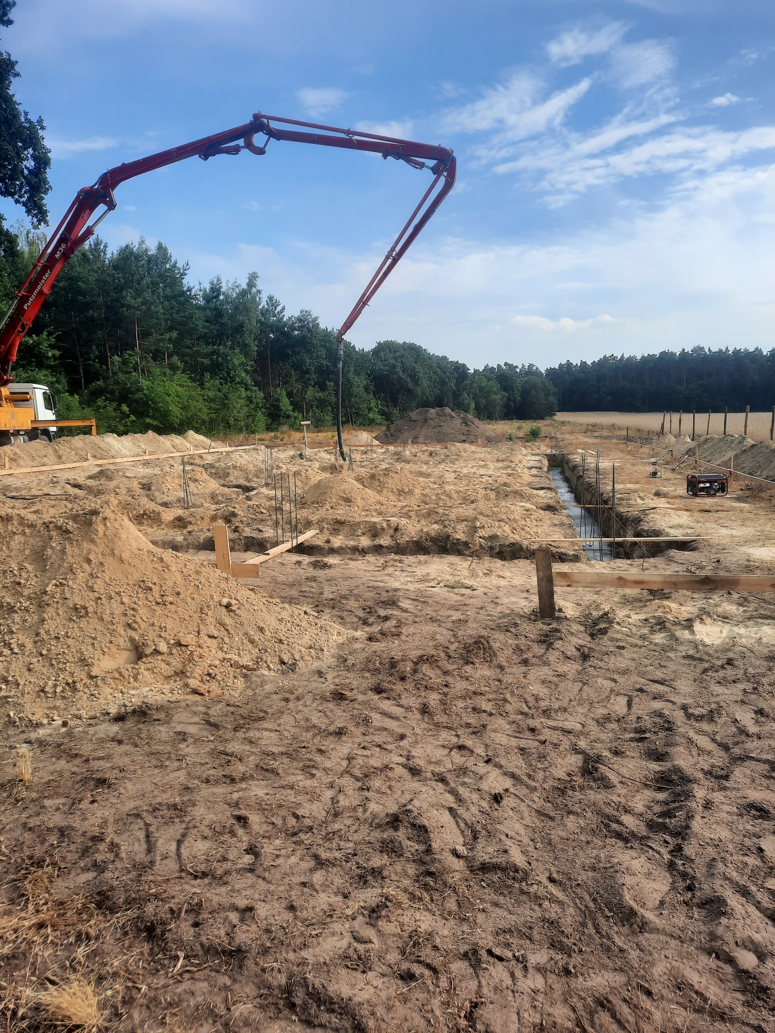 Przygotowanie terenu pod budowę domu murowanego: wykopane fundamenty zbrojone stalą, szalunki z desek, pompa do betonu Putzmeister M36 w tle, teren wyrównany, widok na las w oddali.