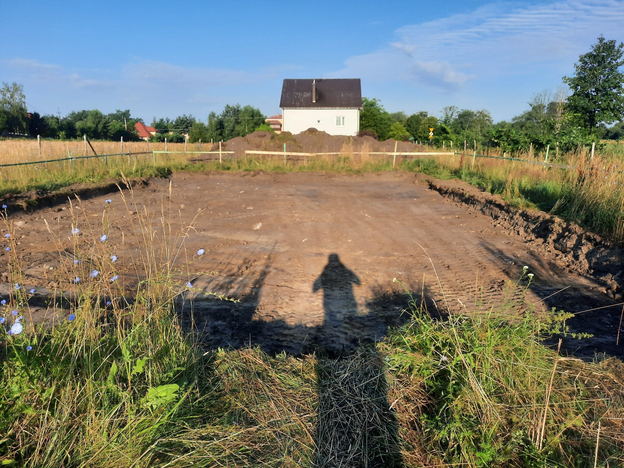 Przygotowany teren pod budowę domu, widoczny fundament, w tle biały dom z ciemnym dachem, cień fotografa na pierwszym planie.