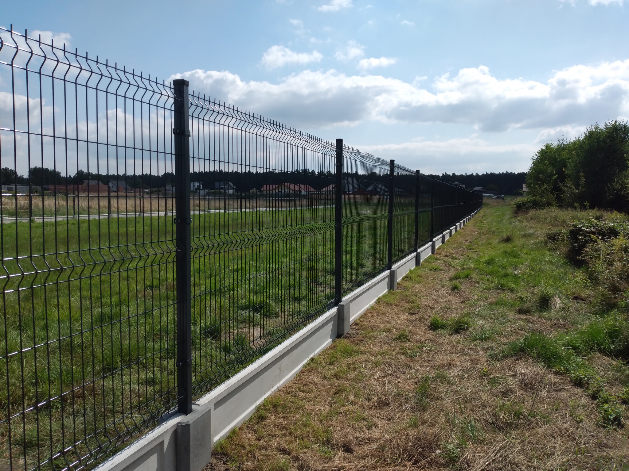 Długie, czarne ogrodzenie panelowe z podmurówką betonową, rozciągające się wzdłuż granicy działki, z widokiem na łąkę i zabudowania w tle pod błękitnym niebem z chmurami.
