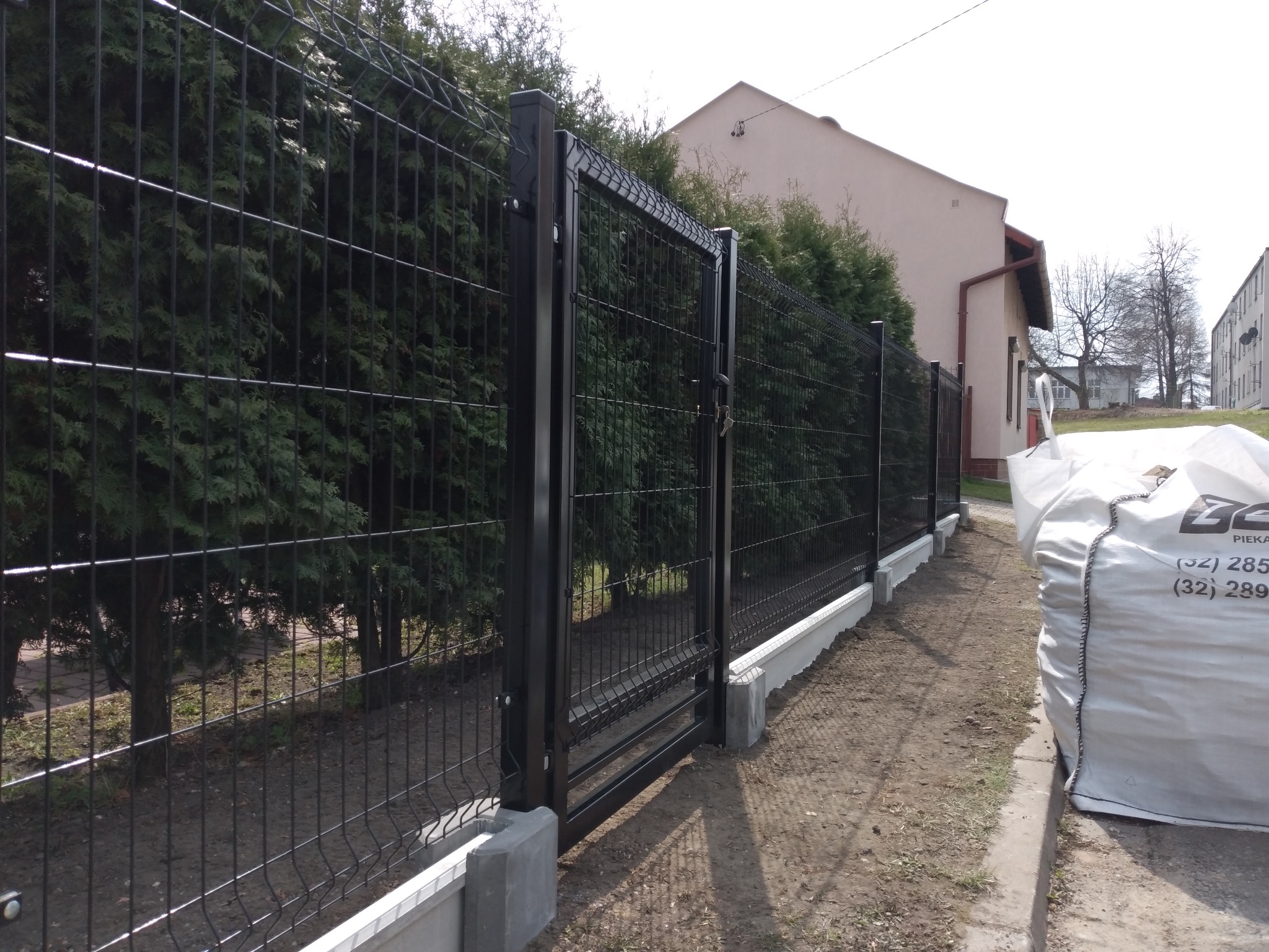 Nowoczesne, czarne ogrodzenie panelowe z bramą wjazdową, osadzone na betonowej podmurówce, z widocznymi zielonymi tujami w tle i workiem z logo firmy po prawej stronie.
