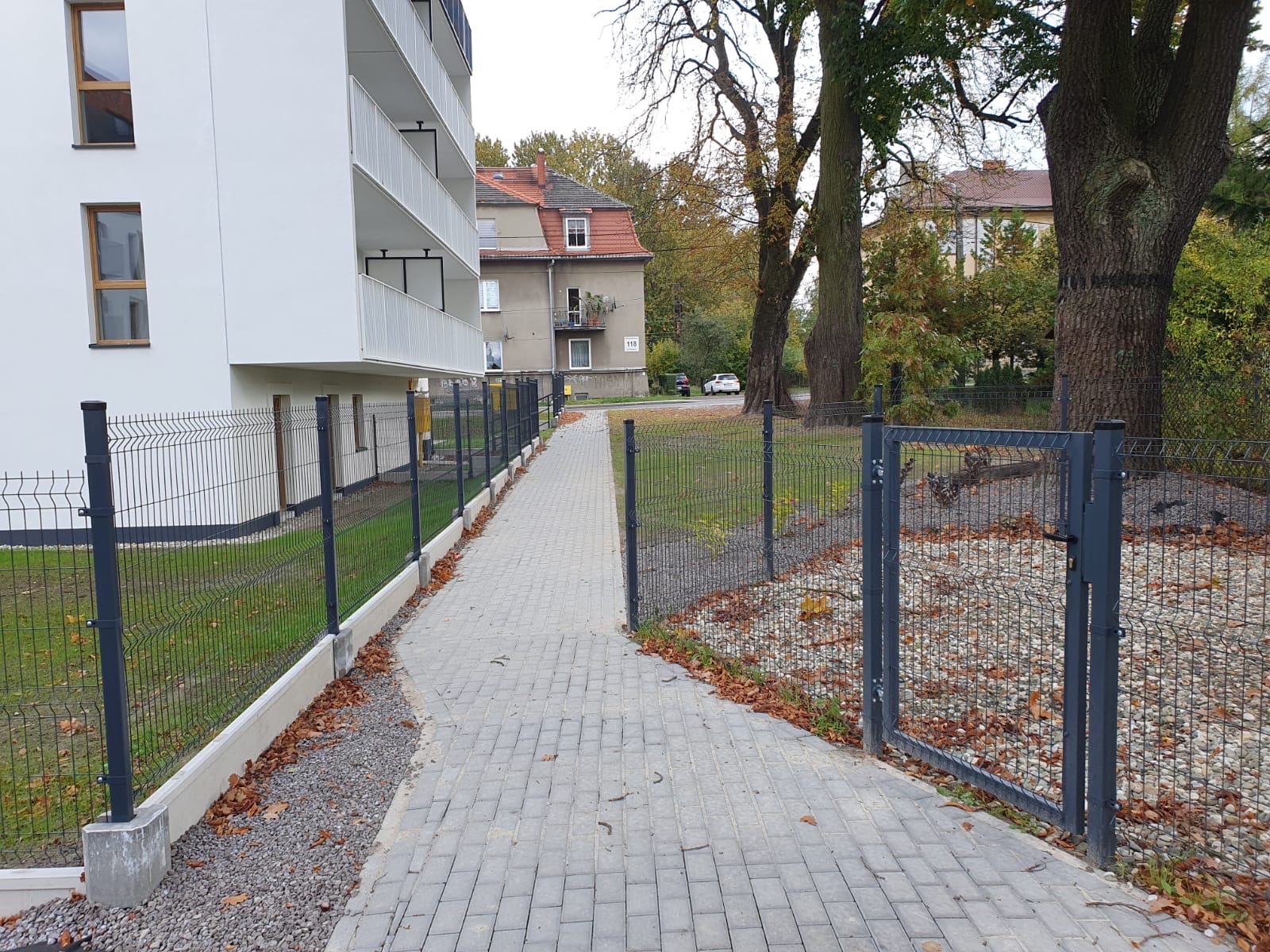 Nowoczesne ogrodzenie panelowe w kolorze grafitowym, oddzielające budynek mieszkalny od chodnika z kostki brukowej i terenu zielonego z drzewami, widoczne jesienne liście.