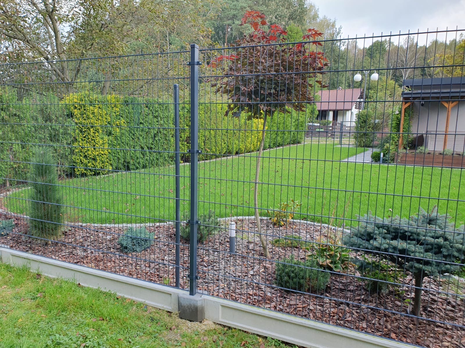 Szary panelowy system ogrodzeniowy z betonową podmurówką otaczający zadbany trawnik z ozdobnymi krzewami i drzewami, widok z zewnątrz posesji.