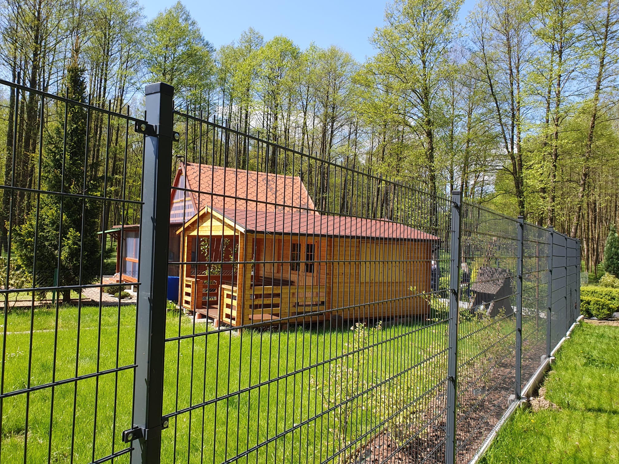 Czarny panelowy płot z widocznym drewnianym domkiem z czerwonym dachem na tle zielonego ogrodu i drzew w słoneczny dzień.