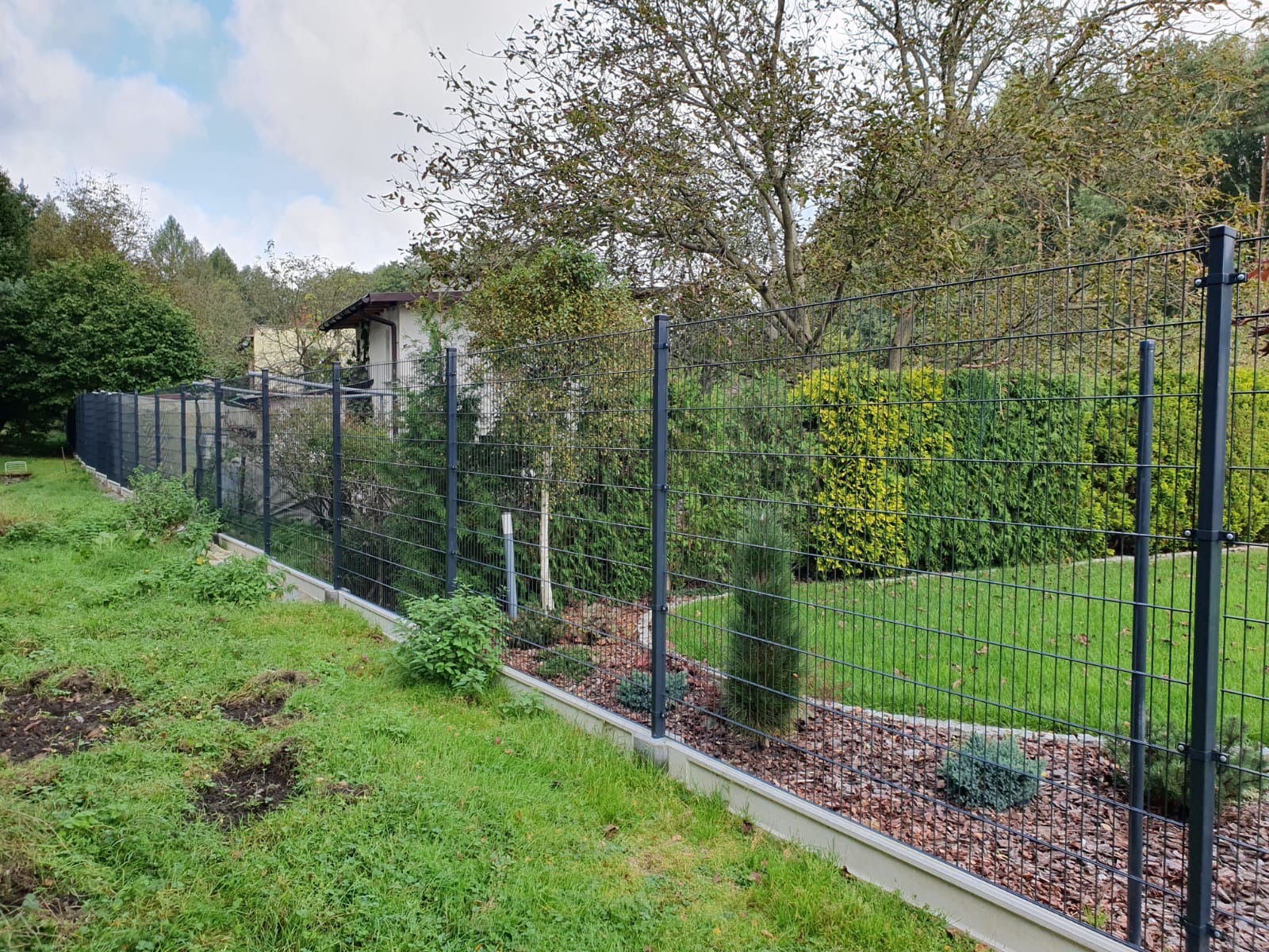 Długie, panelowe ogrodzenie w kolorze grafitowym, oddzielające trawnik od ogrodu z krzewami i drzewami, z betonową podmurówką.