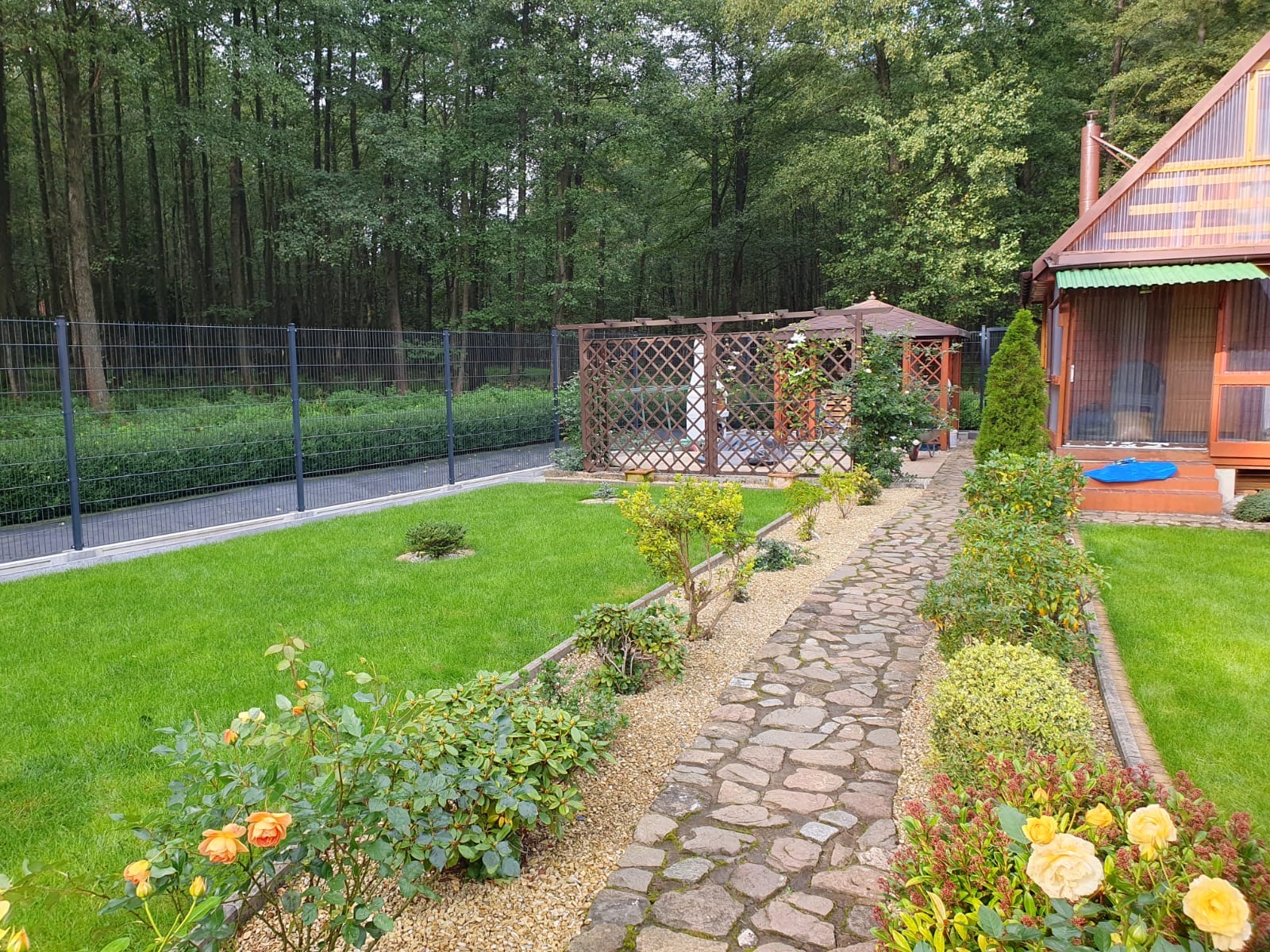 Ogród z szarą siatką ogrodzeniową, drewnianą pergolą i kamienną ścieżką prowadzącą do altany, otoczony zielenią i kwiatami.