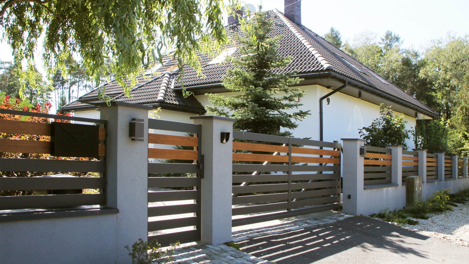 Nowoczesne ogrodzenie z poziomych, szarych elementów metalowych i drewnianych desek, osadzone na szarych, betonowych słupkach z oświetleniem, na tle domu z ciemną dachówką i iglastego drzewa.