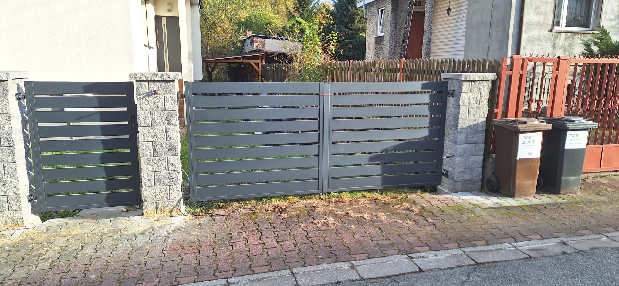 Nowoczesna, szara brama przesuwna z poziomych profili, zamontowana między słupkami z szarej kostki, na tle domów i ogrodów sąsiadów, z widocznymi dwoma pojemnikami na śmieci po prawej stronie.