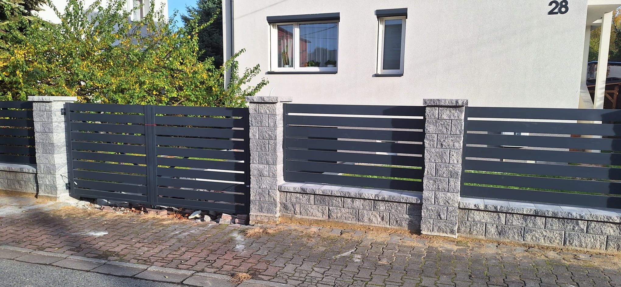 Nowoczesne, poziome przęsła w kolorze grafitowym, osadzone na słupkach z szarej kostki łupanej, tworzące ogrodzenie posesji z numerem 28.