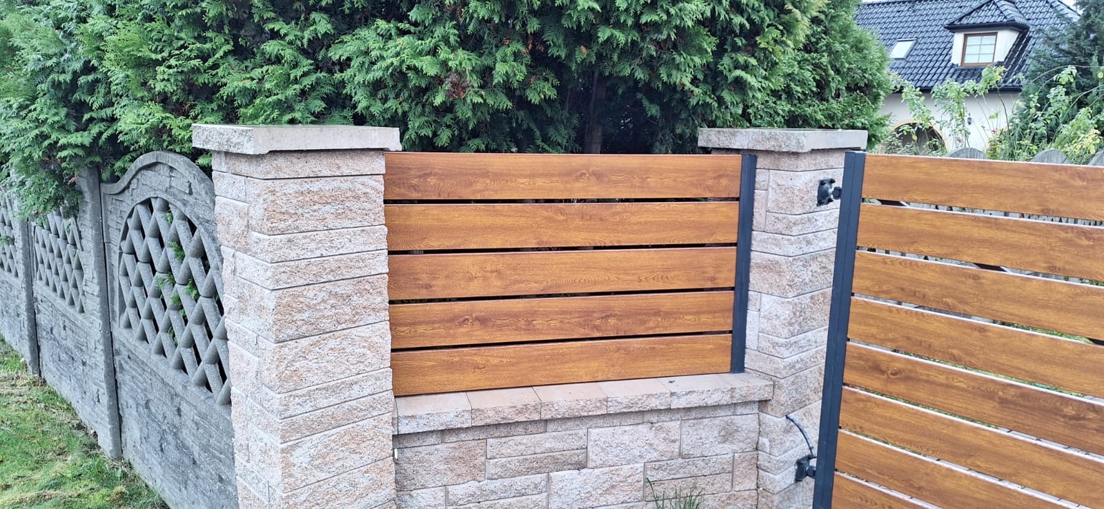 Fragment ogrodzenia z poziomych desek w kolorze drewna, osadzonych między słupkami z jasnego, łupanego kamienia; w tle zielone tuje i dom z ciemną dachówką.