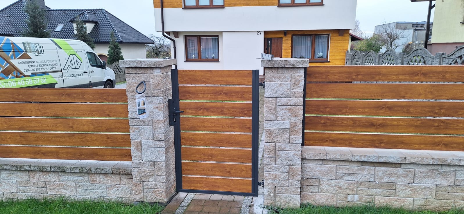 Nowoczesna brama wejściowa z poziomych paneli w kolorze drewna, osadzona w kamiennych słupkach i podmurówce, z widocznym fragmentem domu i samochodu dostawczego firmy w tle.