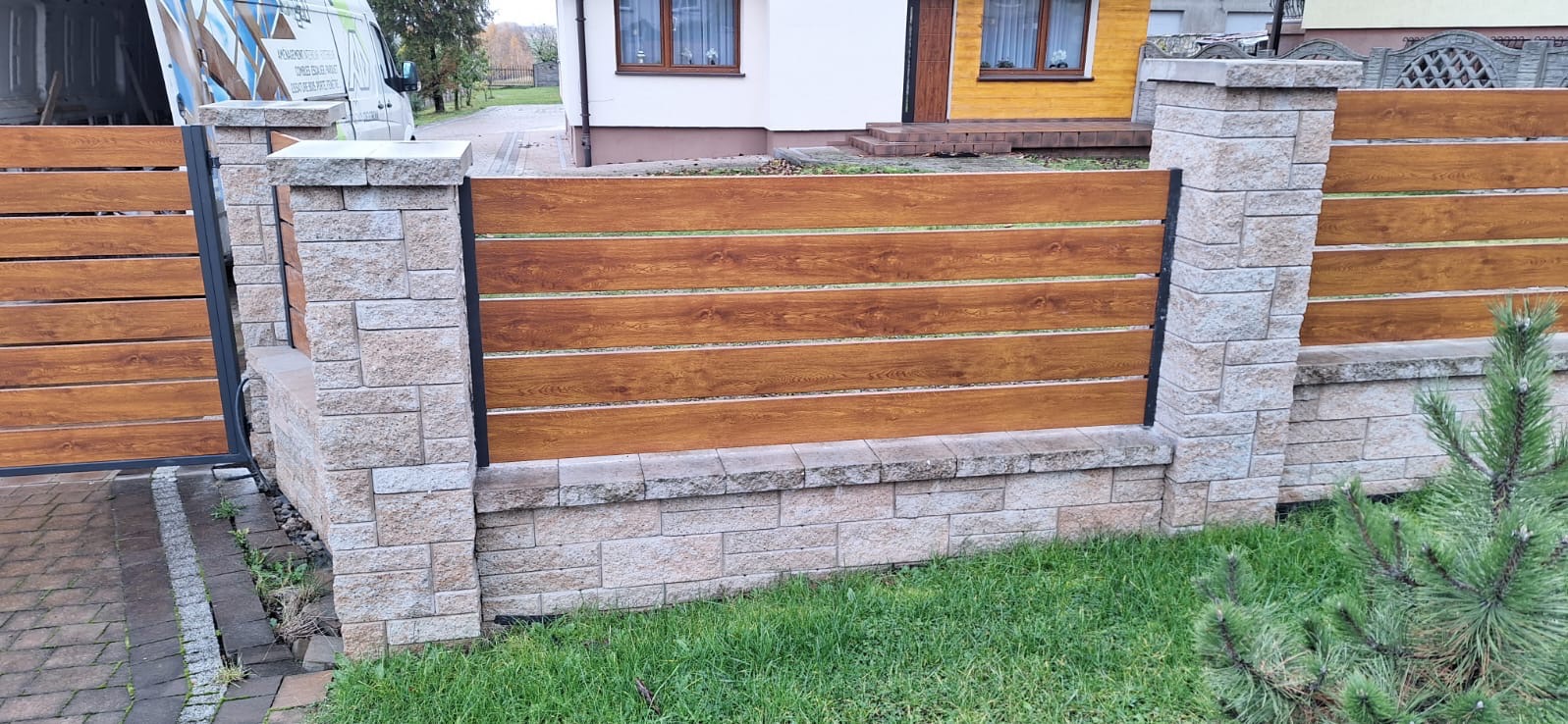 Nowoczesne ogrodzenie z poziomych paneli imitujących drewno, osadzone na słupkach z jasnego kamienia łupanego i czarnych metalowych profilach, z bramą uchylną w tle i fragmentem budynku mieszkalnego.