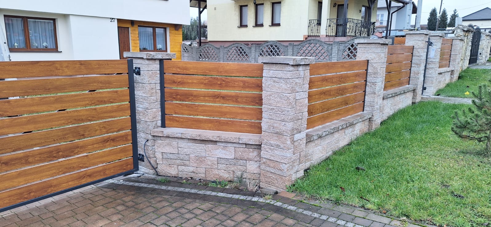 Nowoczesne ogrodzenie posesji z drewnopodobnych paneli w brązowym odcieniu, osadzone na słupkach z jasnego kamienia łupanego, z furtką na brukowanym podjeździe.