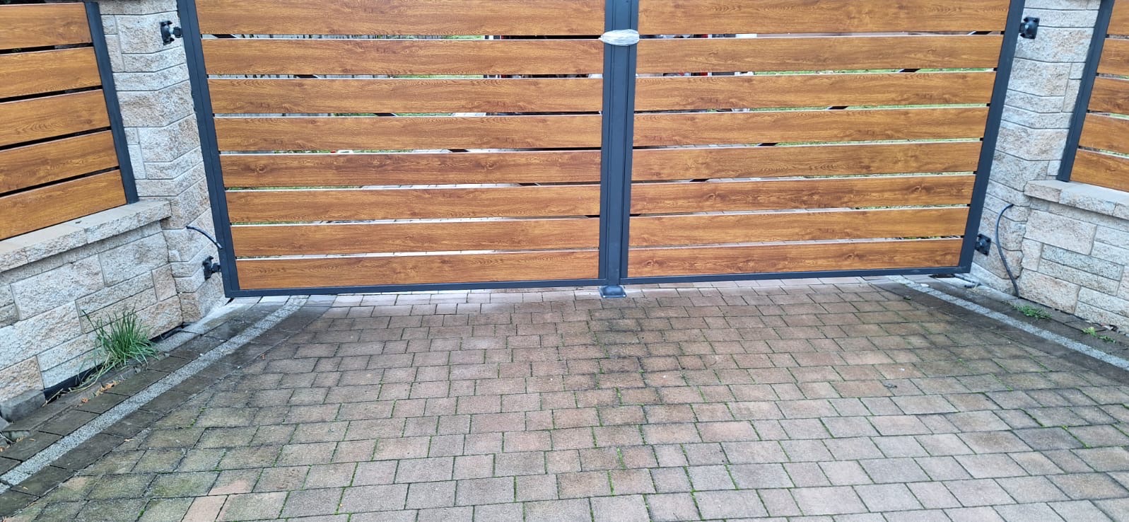 Dwuskrzydłowa brama wjazdowa z poziomych desek imitujących drewno, osadzona na słupkach z szarego kamienia i czarnej metalowej ramie, widok z zewnątrz na kostkę brukową.