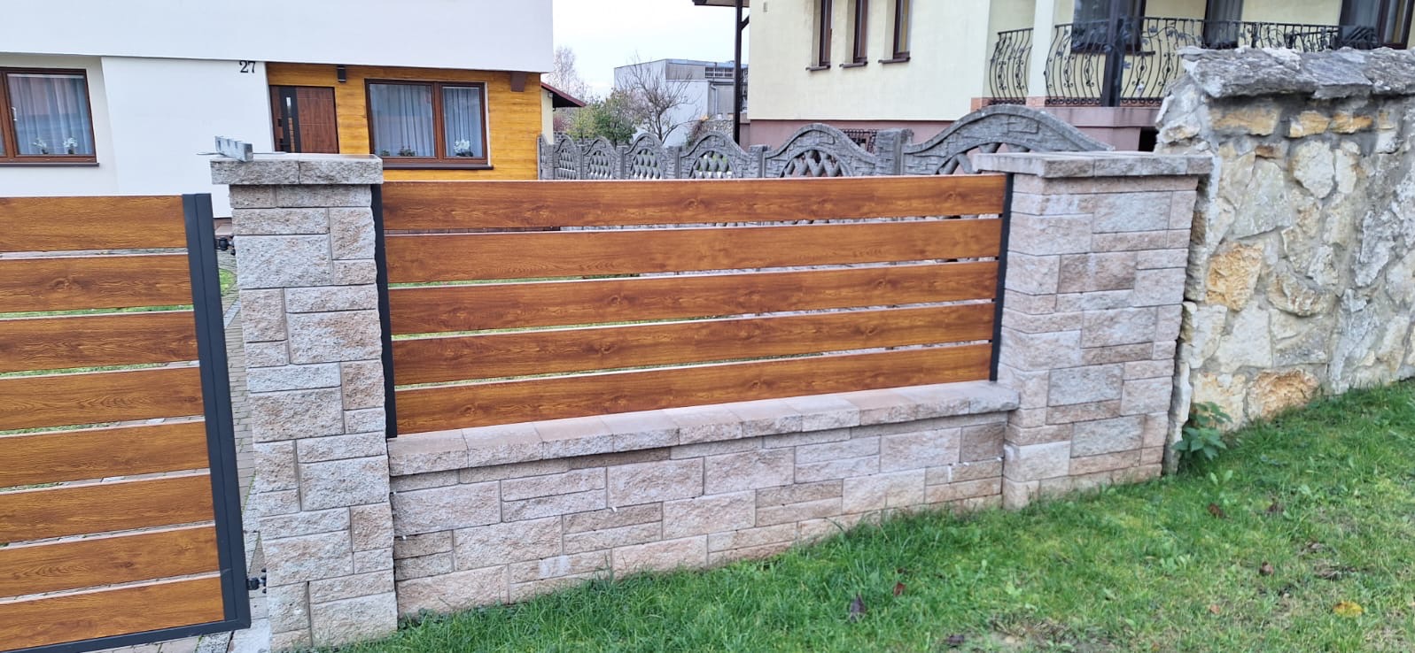 Fragment ogrodzenia z poziomych desek o fakturze drewna, osadzonych w słupkach i podmurówce z jasnego kamienia łupanego, na tle domu z żółtą elewacją i betonowego ogrodzenia sąsiada.