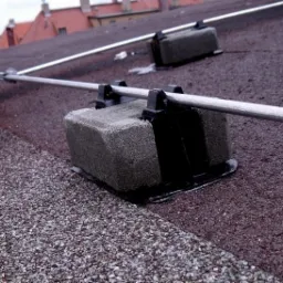 Szara instalacja odgromowa na dachu pokrytym gontem bitumicznym, z widocznymi elementami mocującymi i metalowym przewodem, w tle budynki z czerwonymi dachówkami.