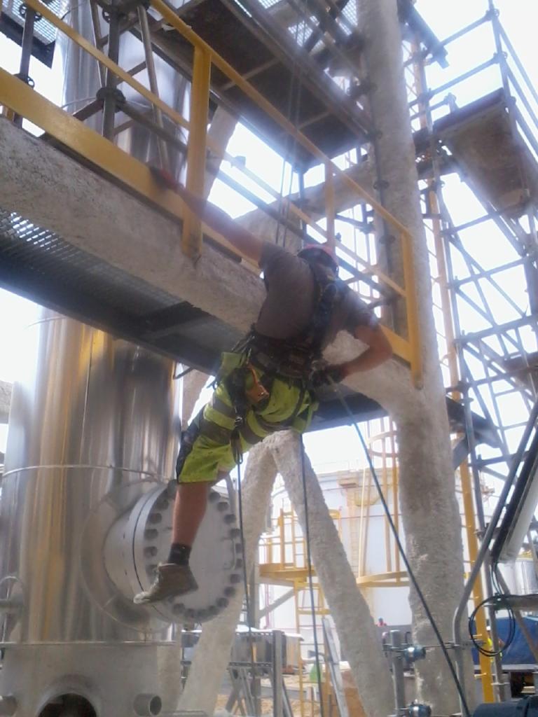 Pracownik w uprzęży alpinistycznej, w trakcie prac na wysokościach przy instalacji przemysłowej, zabezpieczony linami, widoczne elementy konstrukcji stalowej i rury pokryte izolacją.