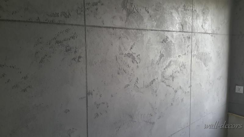 Dekoracyjna, szara ściana z efektem betonu architektonicznego, podzielona pionowymi i poziomymi liniami, tworząc geometryczny wzór. Widoczne nieregularne plamy i przetarcia.