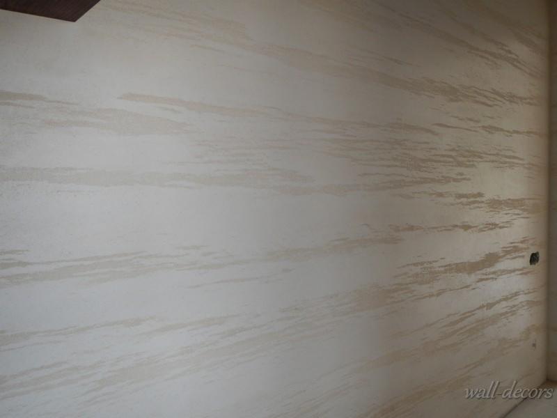 Dekoracyjna ściana w odcieniach beżu i bieli z poziomymi, rozmytymi pasami tworzącymi efekt malarskiego pociągnięcia pędzla. Widoczny narożnik i gniazdko elektryczne.
