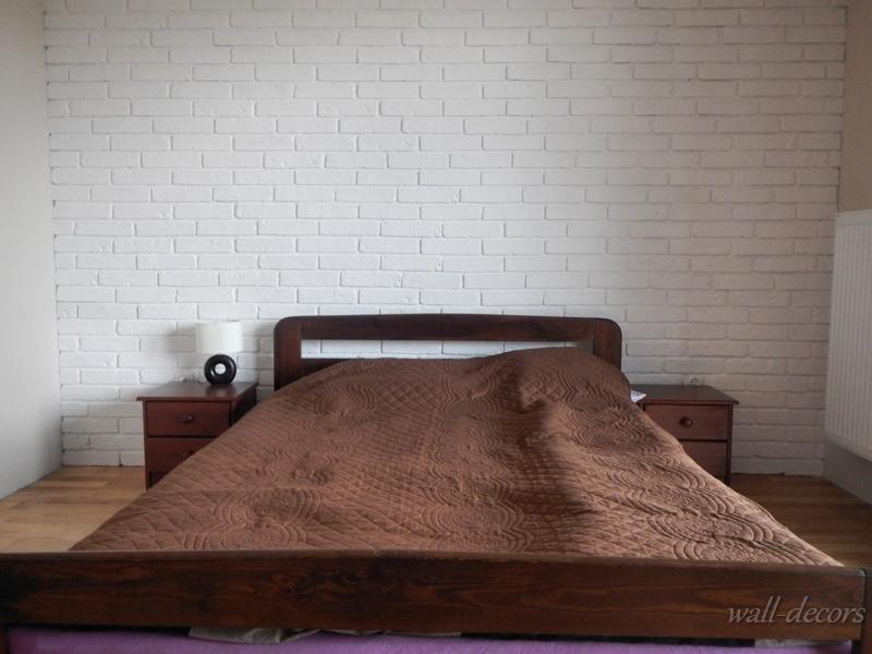 Biała ściana z cegły w sypialni z drewnianym łóżkiem i szafkami nocnymi, lampka na szafce. Widoczny fragment grzejnika. Dekoracja ściany imitująca cegłę.