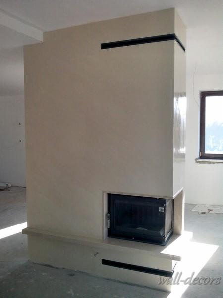 Wykończona obudowa kominka z jasną, gładką powierzchnią. Czarny wkład kominkowy i minimalistyczne detale. Okno w tle. Nowoczesny design.