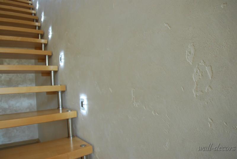 Ściana z dekoracyjną fakturą w odcieniach beżu, oświetlona punktowymi lampami. Obok drewniane schody ze stalowymi elementami, widoczne z boku.
