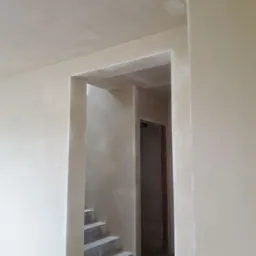 Wnętrze po nałożeniu gładzi gipsowej, widoczne fragmenty schodów i ościeżnica drzwiowa, surowy stan wykończenia.