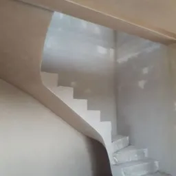 Klatka schodowa w trakcie tynkowania maszynowego, widoczne surowe schody i świeżo nałożony tynk na ścianach i suficie, z widocznymi niedoskonałościami i przewodami na podłodze.