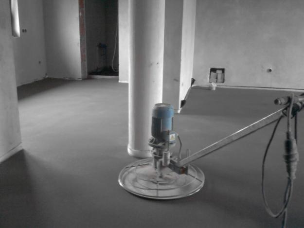 Świeżo wylana i wygładzona posadzka w szarym odcieniu, obok stoi maszyna do zacierania betonu z niebieskim silnikiem, widoczne surowe ściany i filary budynku w tle.