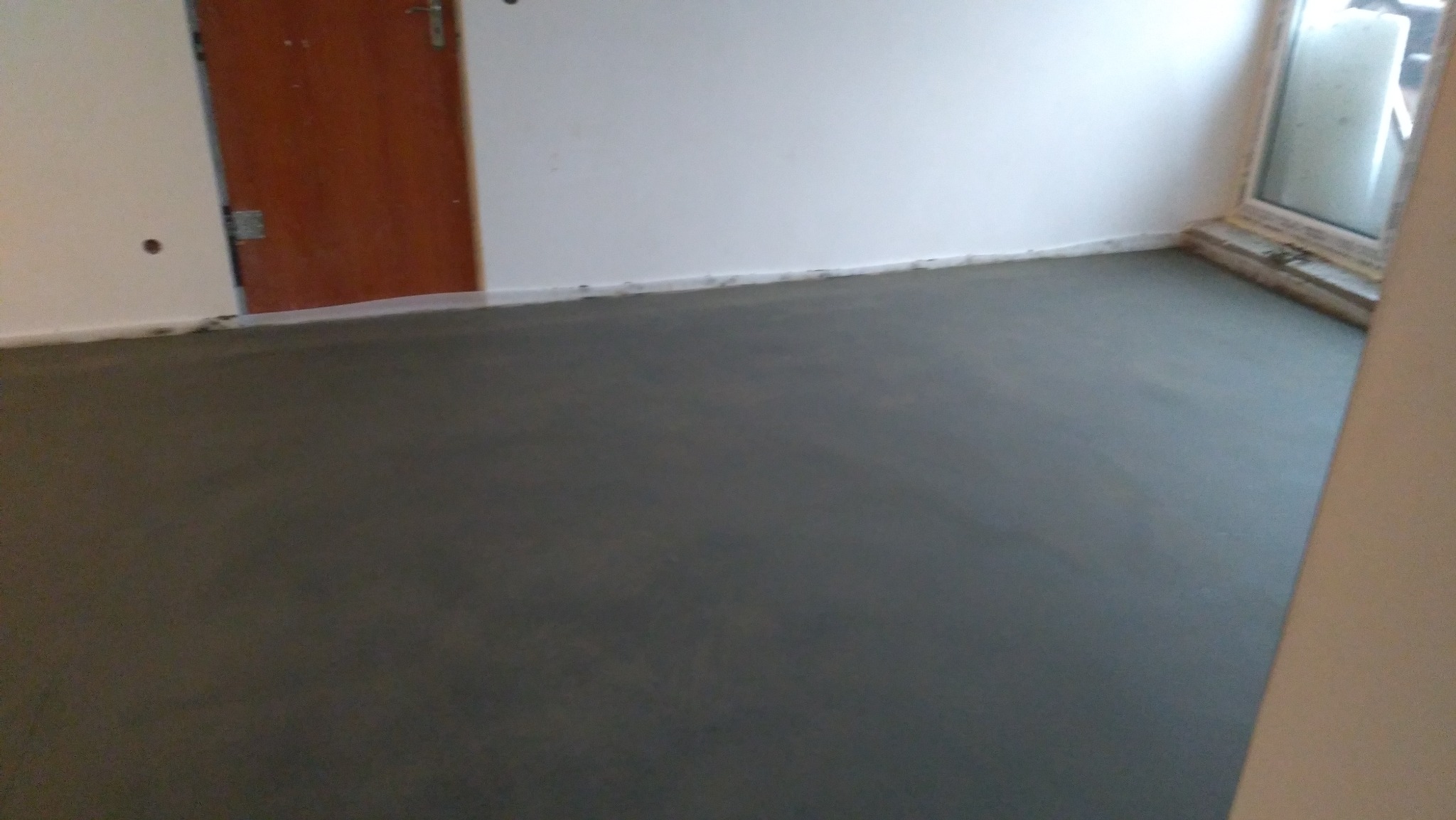 Świeżo wylana, gładka, szara posadzka w pomieszczeniu z białymi ścianami i brązowymi drzwiami, widoczne okno w tle.