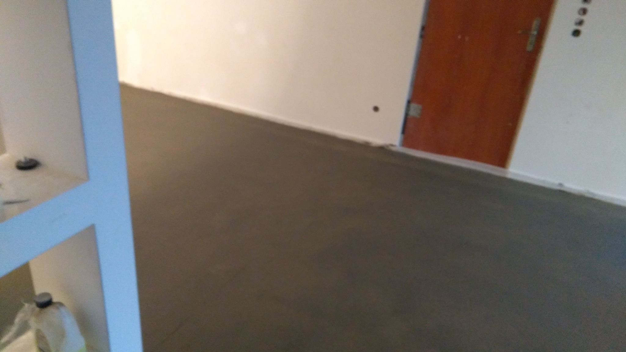 Świeżo wylana, szara posadzka w pomieszczeniu z białymi ścianami i brązowymi drzwiami, widok z góry na gładką powierzchnię.